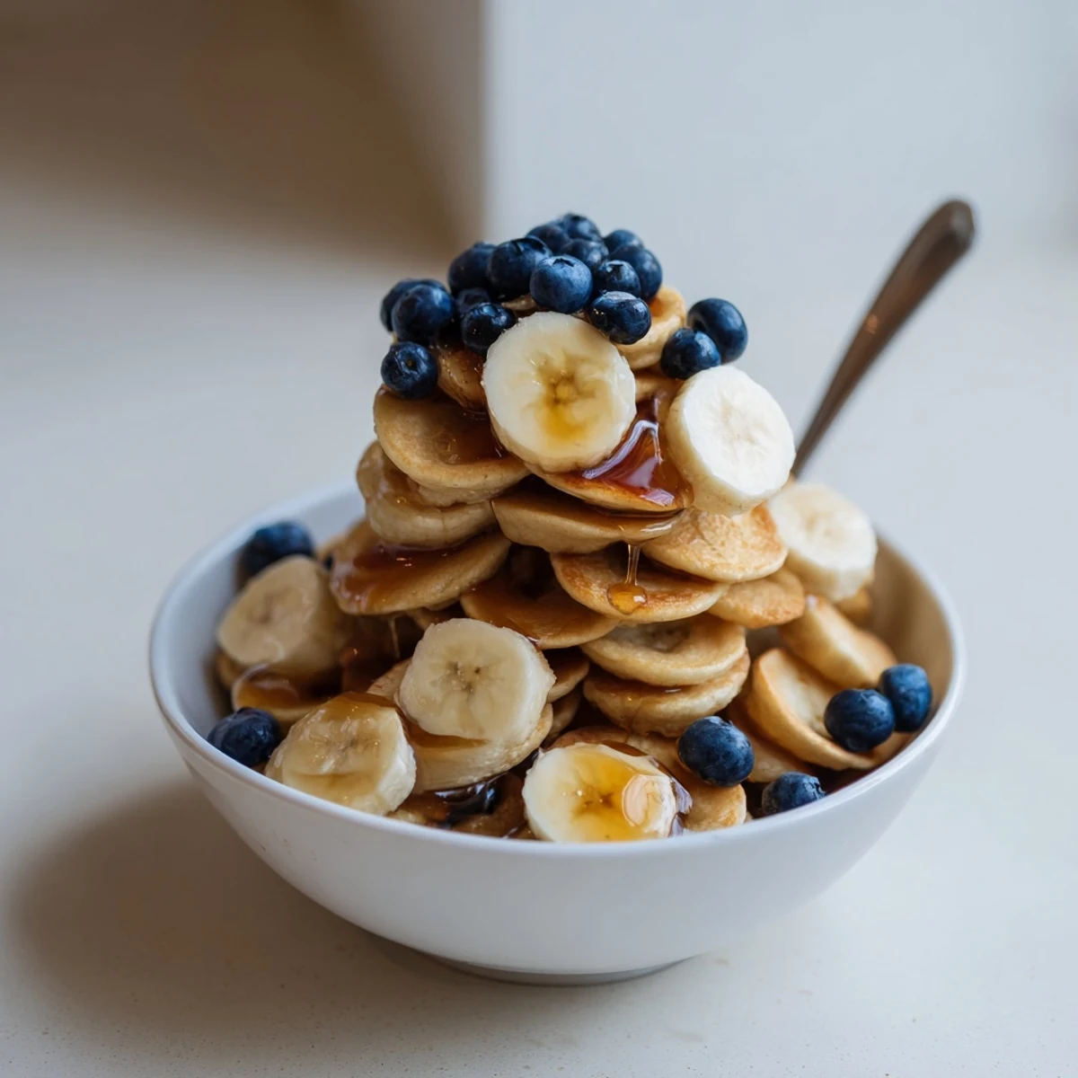 Fluffy mini pancake cereal in a bowl, drizzled with honey and sliced bananas.  